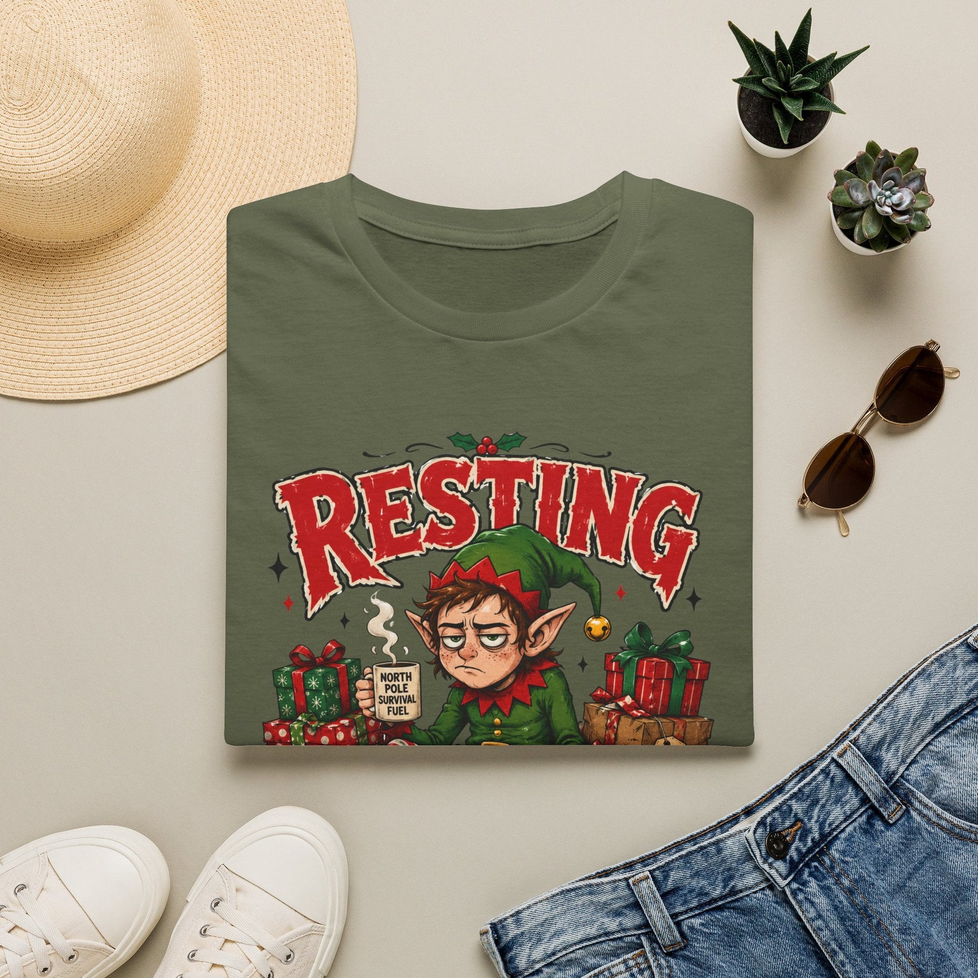 Folded Resting Elf Face Christmas T-Shirt in military green showing a funny Christmas elf graphic design.