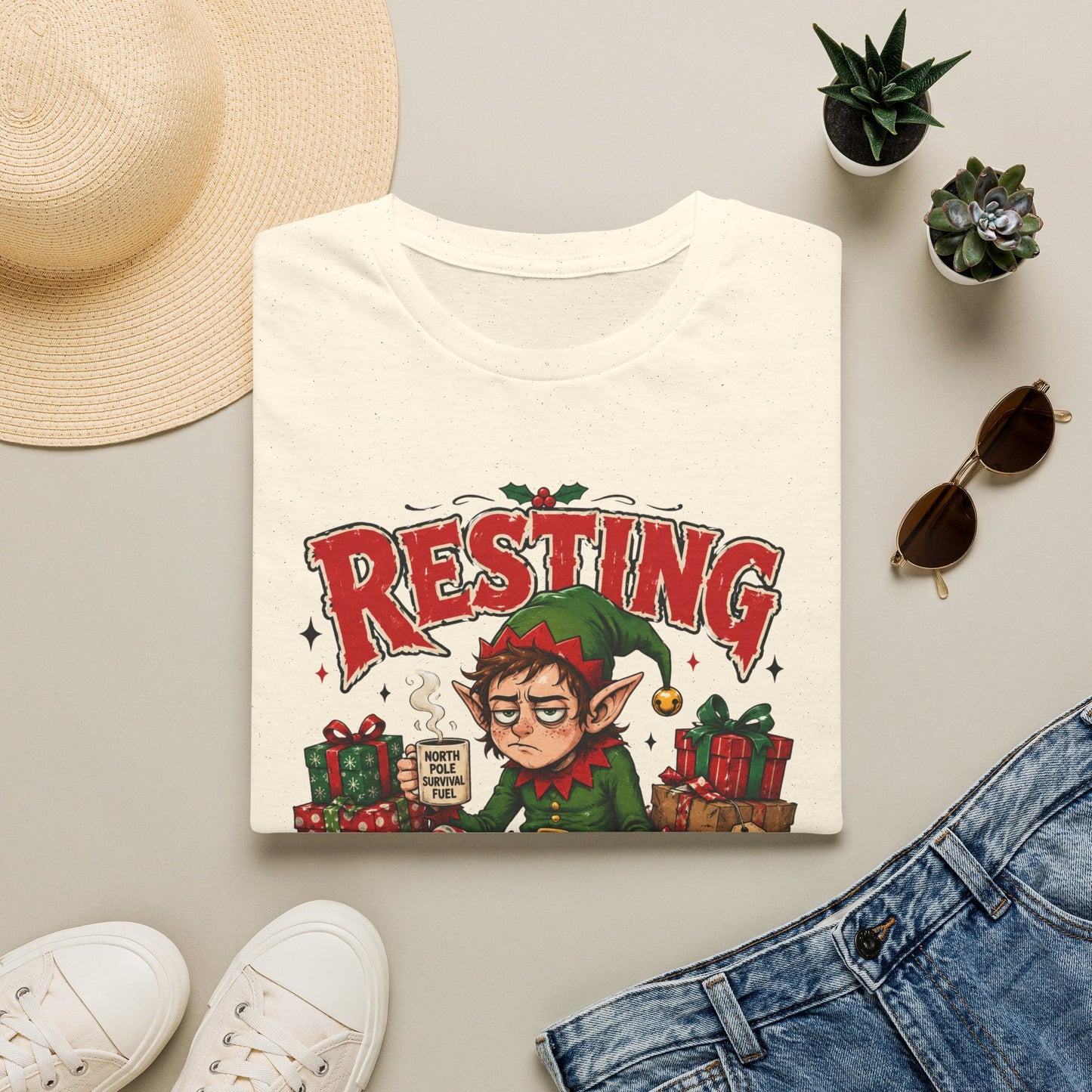Folded Resting Elf Face Christmas T-Shirt in natural showing a funny Christmas elf graphic design.