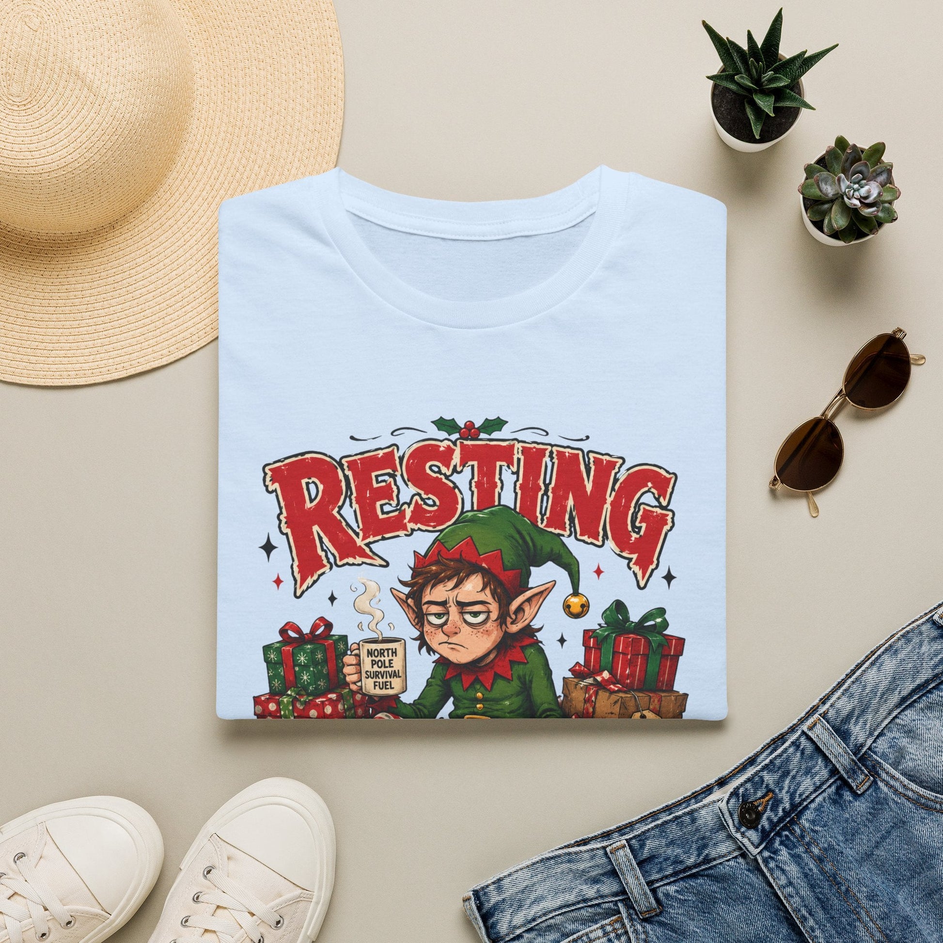 Folded Resting Elf Face Christmas T-Shirt in light blue showing a funny Christmas elf graphic design.